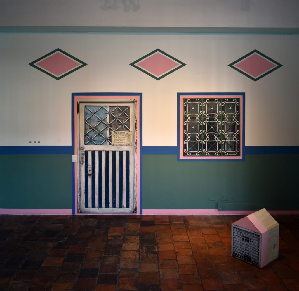 Instalación in situ, Claustro de San Agustín, Bogotá. Puertas, ventanas y elementos domésticos provenientes de viviendas reales se articulan con pintura mural y códigos QR georreferenciados, trasladando fragmentos del territorio al espacio expositivo sin desvincularlos de sus lugares de origen.